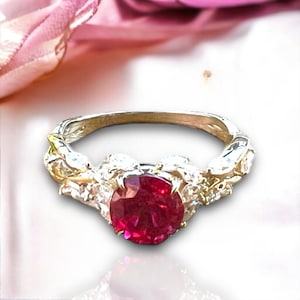 Puede incluir: Un anillo de plata con una gran piedra preciosa de rubí roja redonda rodeada de piedras preciosas blancas más pequeñas.