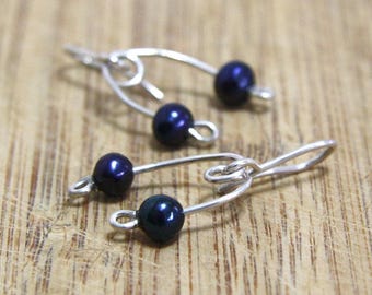 May include: Two pairs of handcrafted earrings. Each earring features a dark blue pearl bead suspended from a silver-toned wire. The wire is shaped into a loop and a hook. The earrings are displayed on a wooden surface.