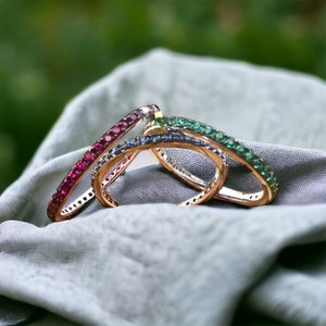 May include: Three gold rings with colourful gemstones. The rings are stacked on top of each other. The top ring has green gemstones, the middle ring has blue gemstones, and the bottom ring has red gemstones.