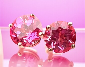 May include: A pair of stud earrings featuring large, faceted, pink gemstones. The gems are set in gold-colored prongs, and the earrings are displayed against a soft pink background, creating a glamorous and eye-catching look. The earrings are round and have a reflective surface.