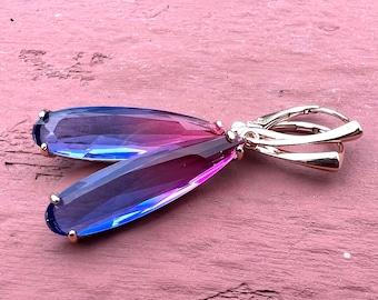 May include: A pair of dangle earrings with a teardrop-shaped, iridescent gemstone that transitions from blue to purple to pink. The earrings are made of gold-toned metal.