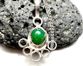 May include: A silver pendant necklace featuring a vibrant green gemstone. The pendant has a unique design with a central stone surrounded by a decorative silver frame and circular accents. The necklace chain is also silver.