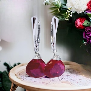 May include: A pair of dangle earrings with faceted, teardrop-shaped, deep red gemstones. The earrings have silver-colored, curved lever-back closures and are accented with silver wire wrapping. The earrings are displayed on a wooden tray with a floral background.