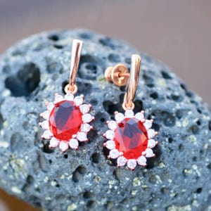 May include: A pair of rose gold earrings featuring oval, deep red gemstones surrounded by small, white gemstones. The earrings have a sunburst design and are displayed on a textured, gray surface.