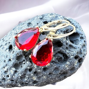 May include: A pair of gold earrings with red teardrop-shaped gemstones. The earrings are on a grey textured surface.
