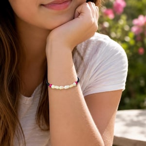 Puede incluir: Una pulsera que presenta una serie de pequeñas cuentas blancas irregulares en el centro, flanqueadas por dos cuentas de color rosa vibrante a cada lado, y completada con segmentos de cordón azul oscuro alrededor de la muñeca.