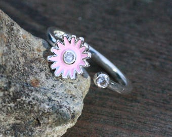 May include: A silver-toned ring featuring a pink daisy flower with a clear center stone and a smaller clear stone on the band. The ring is set against a natural stone and wooden background.