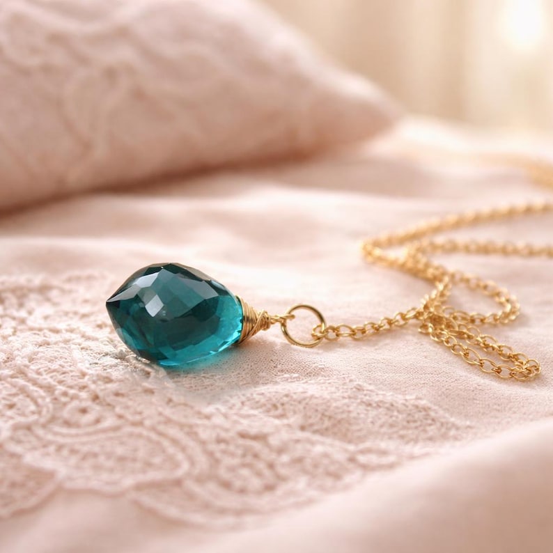 K&ouml;nnte beinhalten: Eine goldene Halskette mit einem gro&szlig;en, facettierten, teal-farbenen Tropfenanh&auml;nger. Der Anh&auml;nger ist mit Golddraht umwickelt. Die Halskette liegt auf einer weichen, rosa, spitzenstrukturierten Oberfl&auml;che.