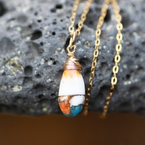 Puede incluir: Un collar dorado con un colgante en forma de lágrima. El colgante presenta una piedra blanca, naranja y turquesa, envuelta en alambre dorado. La fina cadena cuelga sobre un fondo gris texturizado.