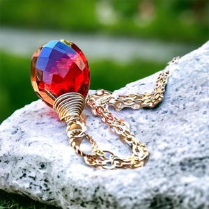 May include: A gold necklace featuring a faceted, teardrop-shaped red and blue gemstone pendant. The gemstone is encased in gold wire and suspended from a delicate gold chain. The necklace is displayed on a textured, grey stone, with a blurred green backdrop.
