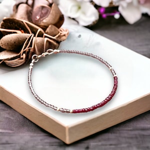 Könnte beinhalten: Ein filigranes Armband mit kleinen, runden Perlen in Lila- und Dunkelrottönen. Das Armband hat einen silbernen Verschluss und wird auf einer weißen Oberfläche präsentiert. Die Perlen sind zu einem einfachen, aber eleganten Accessoire aufgereiht.