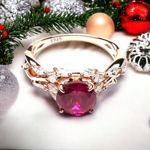 Peut inclure: Une bague en or rose avec une grosse pierre précieuse rubis entourée de petites pierres blanches. La bague a une bande torsadée et l'inscription "S925" à l'intérieur.
