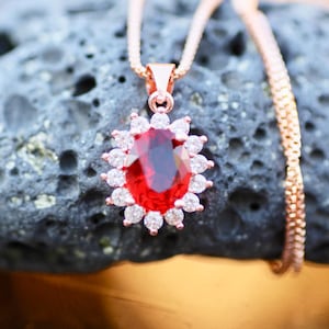 May include: A rose gold necklace with a pendant. The pendant has a large, oval, red gemstone encircled by small, clear crystals. The necklace hangs from a delicate chain. The background is dark and textured.