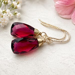 May include: A pair of drop earrings featuring faceted, deep magenta-coloured teardrop stones. Each stone is secured with gold-coloured wire wrapping at the top, connected to textured gold-coloured ear hooks that curve into a simple, elegant shape.