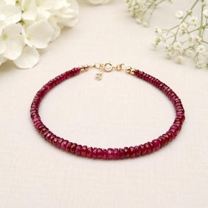 May include: A delicate bracelet composed of small, faceted deep red ruby beads arranged in a continuous circular strand. The piece features a gold-toned metal spring ring clasp and a short extender chain with a small dangling charm at the closure.