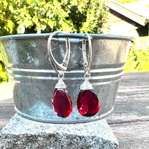 May include: A pair of silver leverback earrings with faceted, teardrop-shaped, deep red gemstones. The gemstones are wire-wrapped at the top and suspended from the silver leverback closures. The earrings are displayed in a galvanized metal bucket.