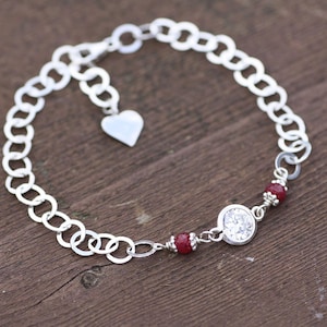 May include: A silver bracelet with a chain link design. The bracelet features a small heart charm, a clear round stone, and two red stones. The bracelet is displayed on a wooden surface.