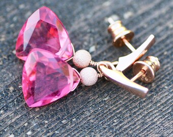 May include: A pair of pink gemstone earrings with a triangular cut. The earrings have a rose gold finish, with a small, textured bead and a post-back closure. The gemstones are a vibrant pink color.