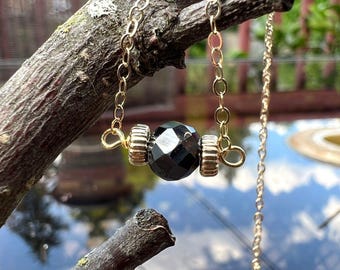 Op de afbeelding: Een goudkleurige ketting met een facetgeslepen zwarte kralen hanger. De hanger wordt geflankeerd door goudkleurige cilindrische kralen. De ketting wordt getoond op een tak, met de achtergrond onscherp.
