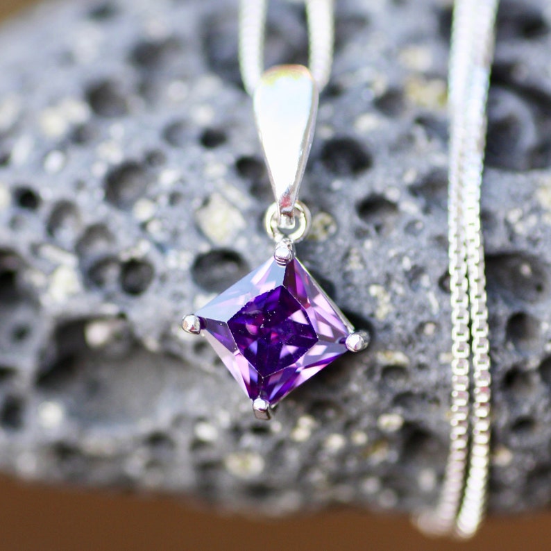May include: A silver pendant necklace with a square-shaped purple gemstone. The pendant is on a silver chain.