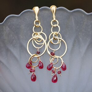 May include: A pair of gold-colored earrings with multiple connected hoops and dangling red gemstones. The earrings have a long, elegant design, with the gemstones adding a pop of color.