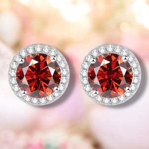 May include: A pair of stud earrings featuring a large, round, faceted, deep red gemstone surrounded by a halo of small, clear, sparkling stones. The earrings have a silver-colored metal setting and are set against a soft, blurred background.