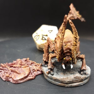 May include: A brown and tan miniature creature with multiple legs and a segmented body, posed on a stone-like base. A separate, similarly colored piece is to the left. A white, multi-sided die is in the background. This is a fantasy game miniature.