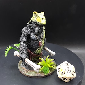 May include: A detailed miniature figurine of a gorilla warrior, wearing a bone helmet and holding bone clubs. The gorilla has a green and brown scale-like armor. A twenty-sided die is next to the figurine.