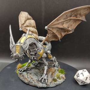 May include: A mechanical dragon figurine with silver and gold metallic plating, featuring large tan leathery wings. The creature stands on a rocky base with small patches of green grass, positioned next to a white twenty-sided die for scale.