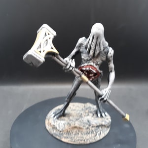 May include: A gray and white monster figurine with a large open mouth and sharp teeth, holding a hammer with a gold-colored handle. The figurine is on a round base.