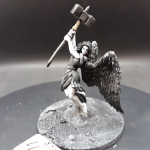 May include: Miniature figurine of an angel with black wings, wielding a hammer. The angel is dressed in a dark gown, with white accents. The figure is standing on a gray, textured base. The hammer is silver and brown.