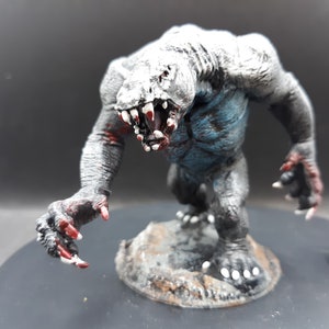 May include: A detailed miniature monster figurine with a menacing expression. The creature has a gray and blue body, sharp white teeth, and red accents. It stands on a small, textured base, suggesting a fantasy or tabletop game theme.