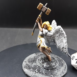 May include: Miniature angel figurine with a hammer raised overhead. The angel has white wings, a white and gold dress, and blonde hair. The figurine is on a gray base, with a white and black die in the background.
