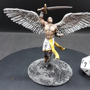 May include: A miniature figurine of an angel with large, detailed wings, holding a sword aloft. The angel is depicted with a muscular physique, wearing a white cloth and a yellow sash. A white die with black numbers is next to the figurine.