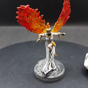 May include: Miniature figurine of a woman with fiery orange and yellow wings, wearing a white dress with gold accents. The figure is on a gray, textured base. A white die with black numbers is in the background.