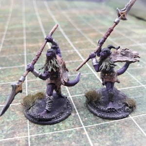 May include: Two miniature fantasy figures, each holding a spear. The figures have purple skin, green beards, and wear brown and tan clothing. They stand on textured, dark bases with patches of faux grass. The spears have silver blades.