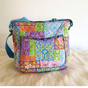 May include: A colorful patchwork crossbody bag with a paisley pattern in various colors, including blue, green, orange, and purple. The bag has a blue adjustable strap and a front pocket. The bag is made of quilted fabric.
