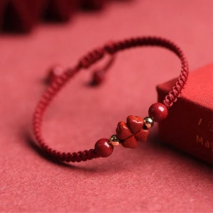 May include: A red string bracelet with a red clover charm and gold accents. The bracelet is tied with a knot and has a tassel.