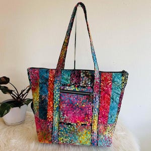 May include: A colorful tote bag with a patchwork design. The bag features a variety of vibrant colors, including red, blue, yellow, and pink. It has a zippered top, exterior pockets, and long handles. The bag is made of quilted fabric.
