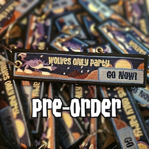 Könnte beinhalten: Gestickter Schlüsselanhänger mit dem Text "Wolves Only Party" und "Go Now!" vor einem Hintergrund mit Sternenhimmel. Der Schlüsselanhänger zeigt eine Wolfsgrafik und ist von ähnlichen Schlüsselanhängern umgeben. Das Wort "Pre-order" wird darunter angezeigt.