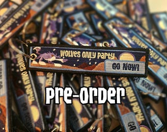 Può includere: Portachiavi ricamato con la scritta "Wolves Only Party" e "Go Now!" su uno sfondo di cielo stellato. Il portachiavi presenta una grafica di lupo ed è circondato da portachiavi simili. La parola "Pre-order" è visualizzata sotto.