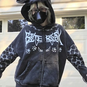 Puede incluir: Una sudadera con capucha con cremallera gris oscuro con una capucha con orejas de oso. La sudadera tiene texto blanco que dice "BITE RISK" y huellas de patas. Las mangas tienen un patrón de diamantes blancos. Se usa una máscara negra.