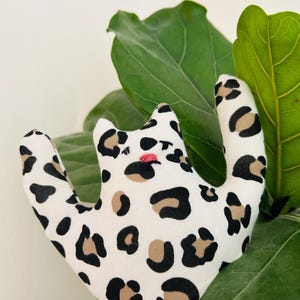 Può includere: Un giocattolo di peluche bianco a forma di gatto con macchie nere e marroni. Il giocattolo ha un naso rosa ed è ambientato su uno sfondo di grandi foglie verdi. Il peluche è un design stravagante e giocoso.