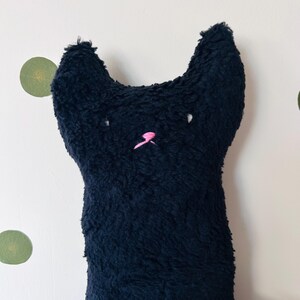 Könnte beinhalten: Ein Plüschtier in Katzenform aus dunkelblauem, strukturiertem Stoff. Die Katze hat zwei spitze Ohren, kleine weiß gestickte Augen sowie eine leuchtend rosa gestickte Nase und Mundpartie in der Mitte des Gesichts.