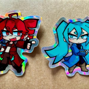 May include: Two holographic stickers featuring anime-style characters. The first sticker is red and features a character with red hair and a red jacket. The second sticker is blue and features a character with blue hair and a blue jacket.