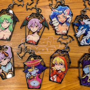 May include: A collection of anime-style character keychains. Each keychain features a different character design within a coffin-shaped frame. The keychains have a silver-colored bat-shaped clasp.