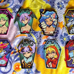 May include: A collection of eight coffin-shaped stickers featuring anime-style illustrations. The stickers showcase various characters with vibrant colors, including green, purple, blue, and red, with bat-shaped accents and a wolf.