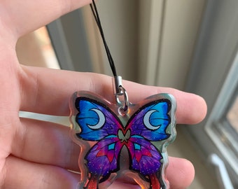 May include: A clear, iridescent butterfly charm with a purple and blue design and two crescent moons on the wings. The charm is attached to a black string.