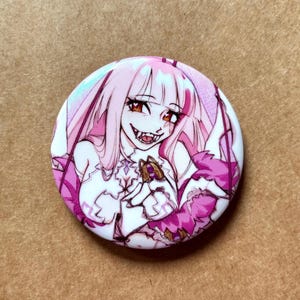 May include: A round button with a cartoon illustration of a character with pink hair, red eyes, and sharp teeth. The character is wearing a white and pink outfit with a floral design. The button has a white background.