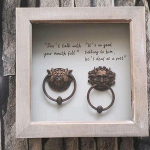 May include: A framed piece of art with two bronze door knockers, each with a detailed face design. The artwork includes the phrases "Don't talk with your mouth full" and "It's no good talking to him, he's deaf as a post."